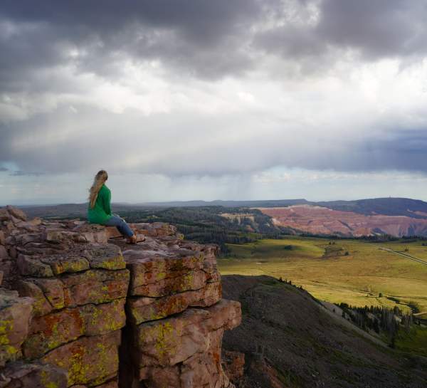 Woman in green jacket sits on the rock outcropping of the Brian Head Peak overlook at 11,000 feet elevation with Cedar Breaks National Monument in the background.