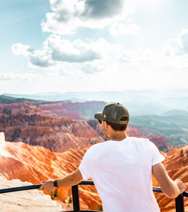 Person standing at scenic overlook at Cedar Breaks National Monument with a red rock canyon in the background and blue skies.