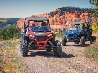 A red OHV followed by a blue OHV round the corner of a dirt road with pine trees and red rock formation in the background.